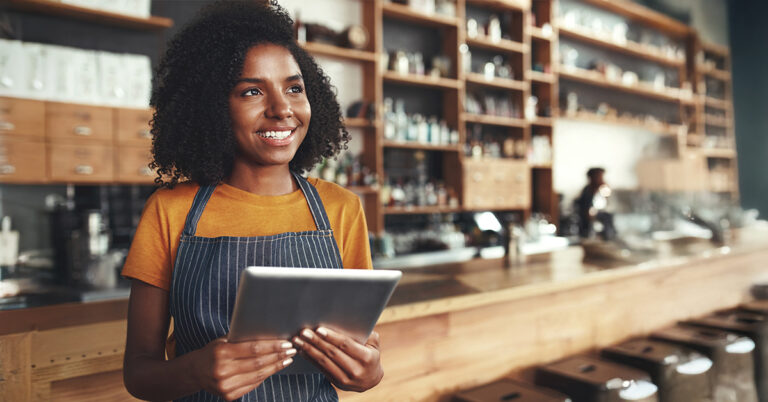 Professional female alcohol server in apron standing at sunlit bar, holding tablet and looking thoughtfully into distance