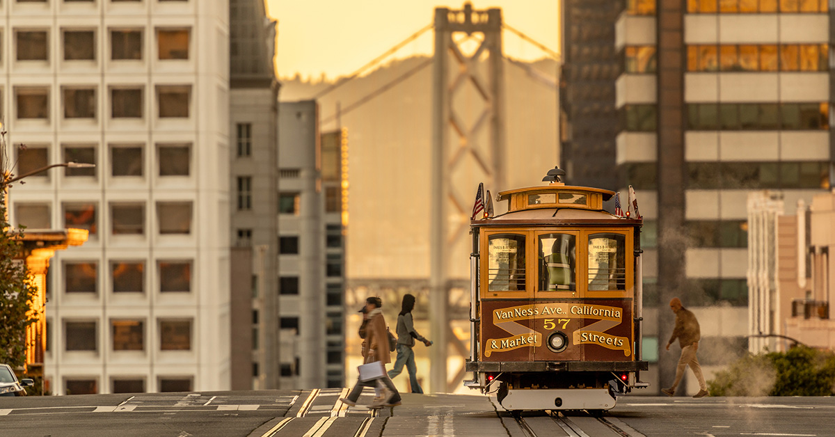 San Francisco street at dusk with pedestrians crossing after a streetcar passes, framed by skyscrapers with the iconic Golden Gate Bridge visible in the distance, capturing California's urban hospitality scene.